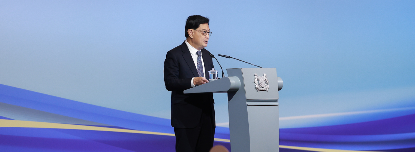 Man in suit speaking at podium with Singapore crest against blue gradient background.
